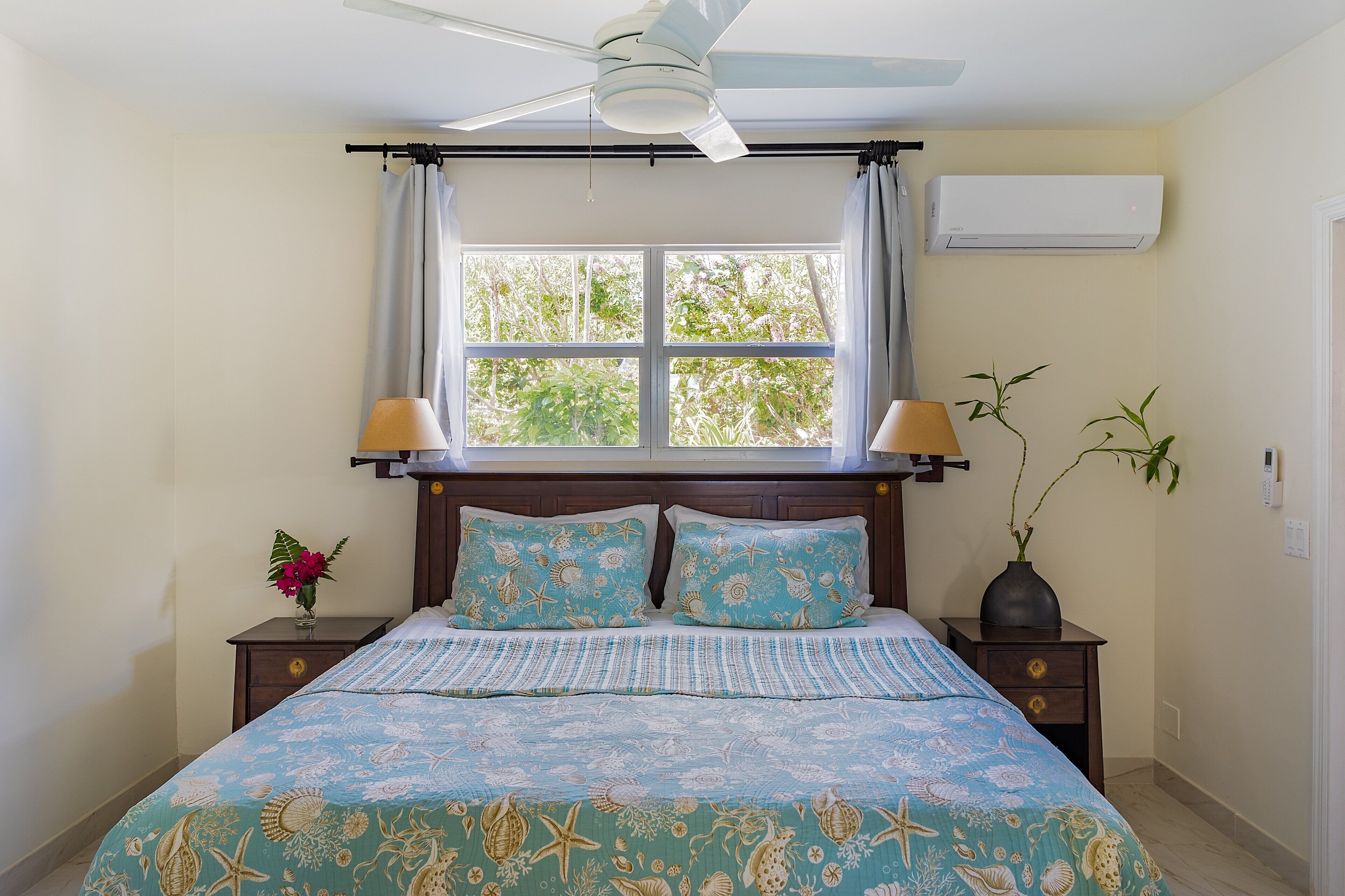 A bedroom with a bed, a ceiling fan, a window with curtains, an air conditioner, a nightstand with a lamp, and a vase with a plant.