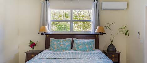 A bedroom with a bed, a ceiling fan, a window with curtains, an air conditioner, a nightstand with a lamp, and a vase with a plant.