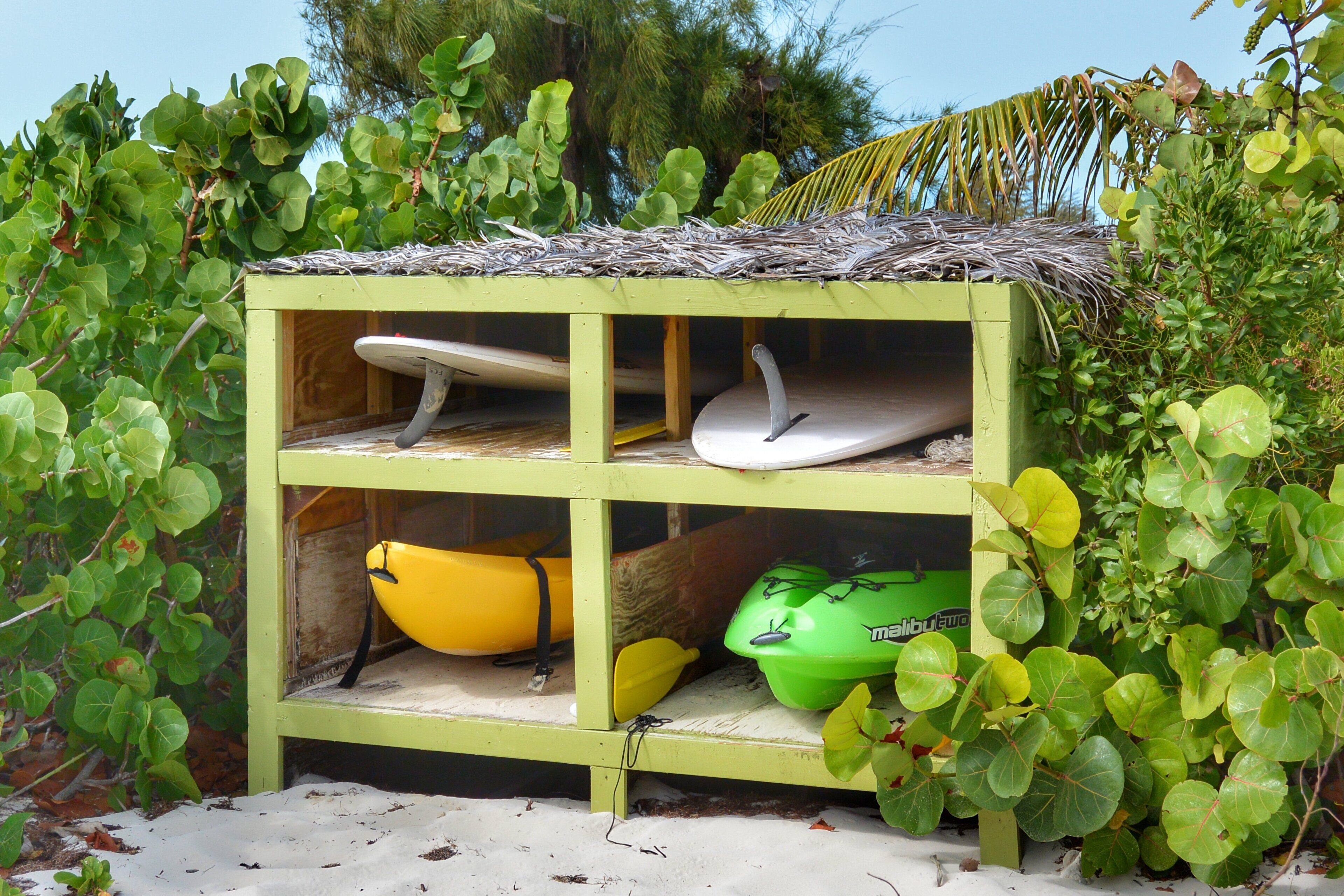 Une petite structure en bois, de couleur verte, avec un toit de chaume, à l’intérieur de laquelle se trouvent une planche de surf blanche, un kayak jaune et un kayak vert.