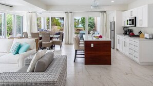 A spacious living area with a sofa, a dining table, and chairs, a kitchen with white cabinets and appliances, and large windows offering a view of greenery.