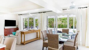A spacious living area with a dining table set for four, surrounded by wicker chairs. A flat-screen TV is mounted on the wall, and the room has large sliding glass doors leading to an outdoor area.