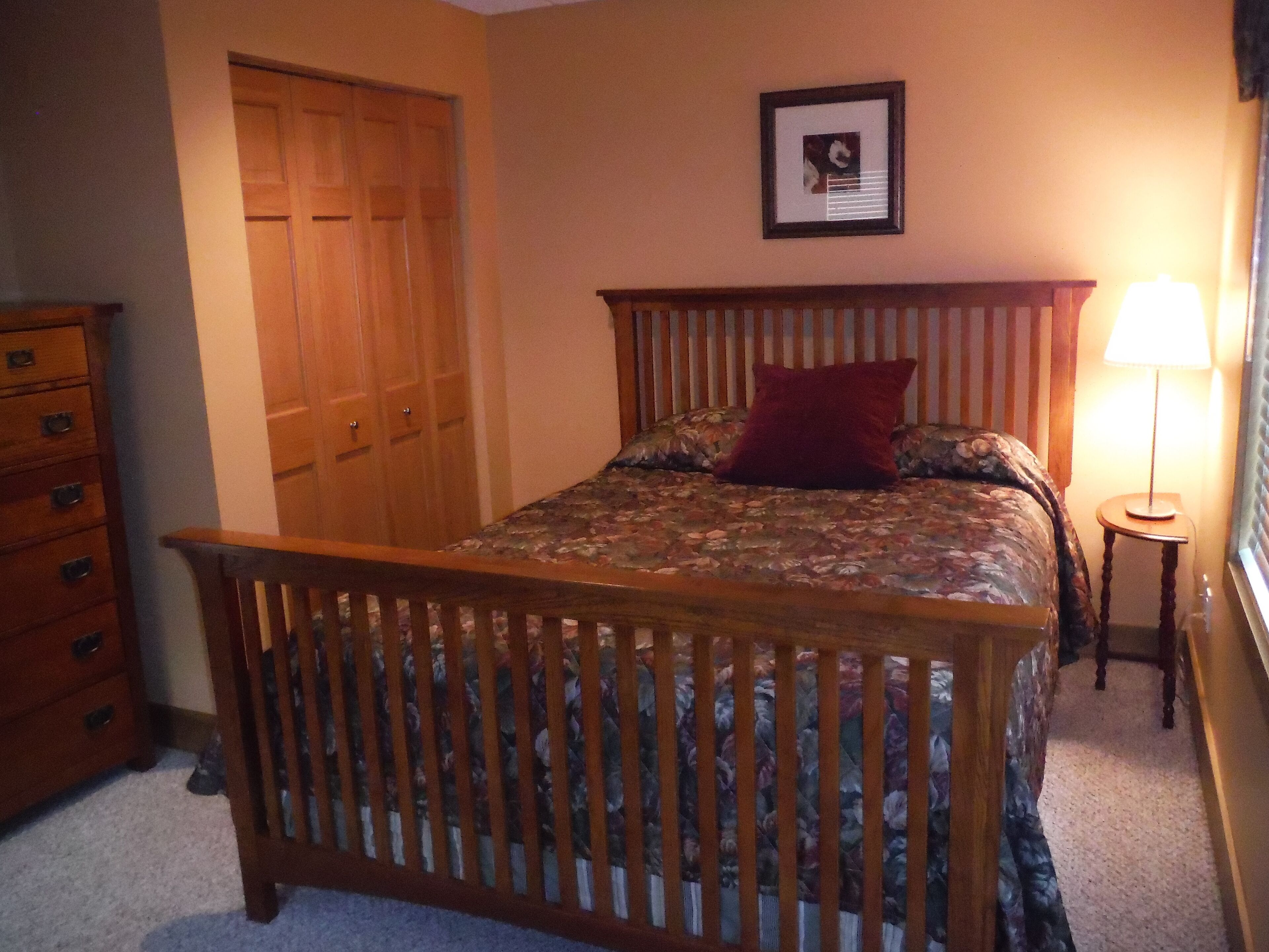 Un dormitorio con estructura de cama de madera, cubrecama floral, almohada roja, cómoda de madera y un cuadro en la pared.