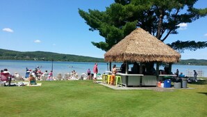 A beachside area with a thatched-roof structure, possibly a bar or kiosk, surrounded by grass and a body of water. - Boyne Mountain on Resort Condo, close to Lifts, Lodge, Waterpark, all amenities (Boyne Falls)