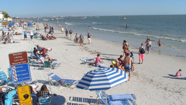Una playa concurrida donde la gente se toma el sol y nada. Hay carteles que indican que no se permiten mascotas, ni alcohol, ni salvavidas.