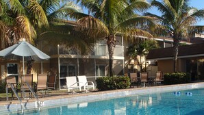 Zona de piscina con sillones de descanso y sombrillas, rodeada de palmeras y un edificio con balcones.
