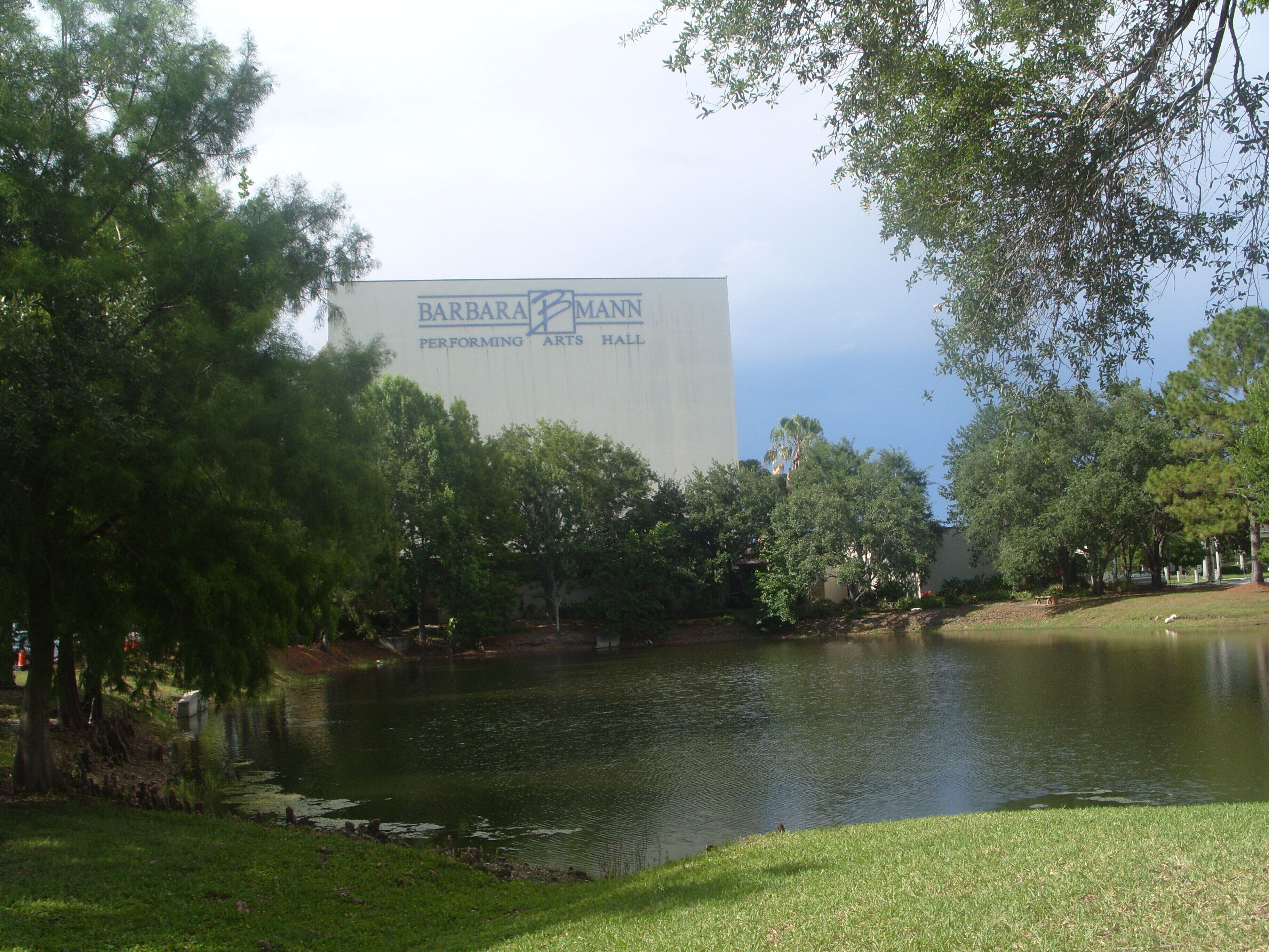 A park with a pond, trees, and a large building with 'BARBARA MANN PERFORMING ARTS HALL' written on it.