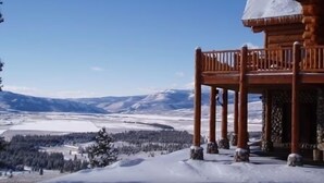 Eine Holzblockhütte mit einem Balkon mit Blick auf eine verschneite Landschaft und ferne Berge.