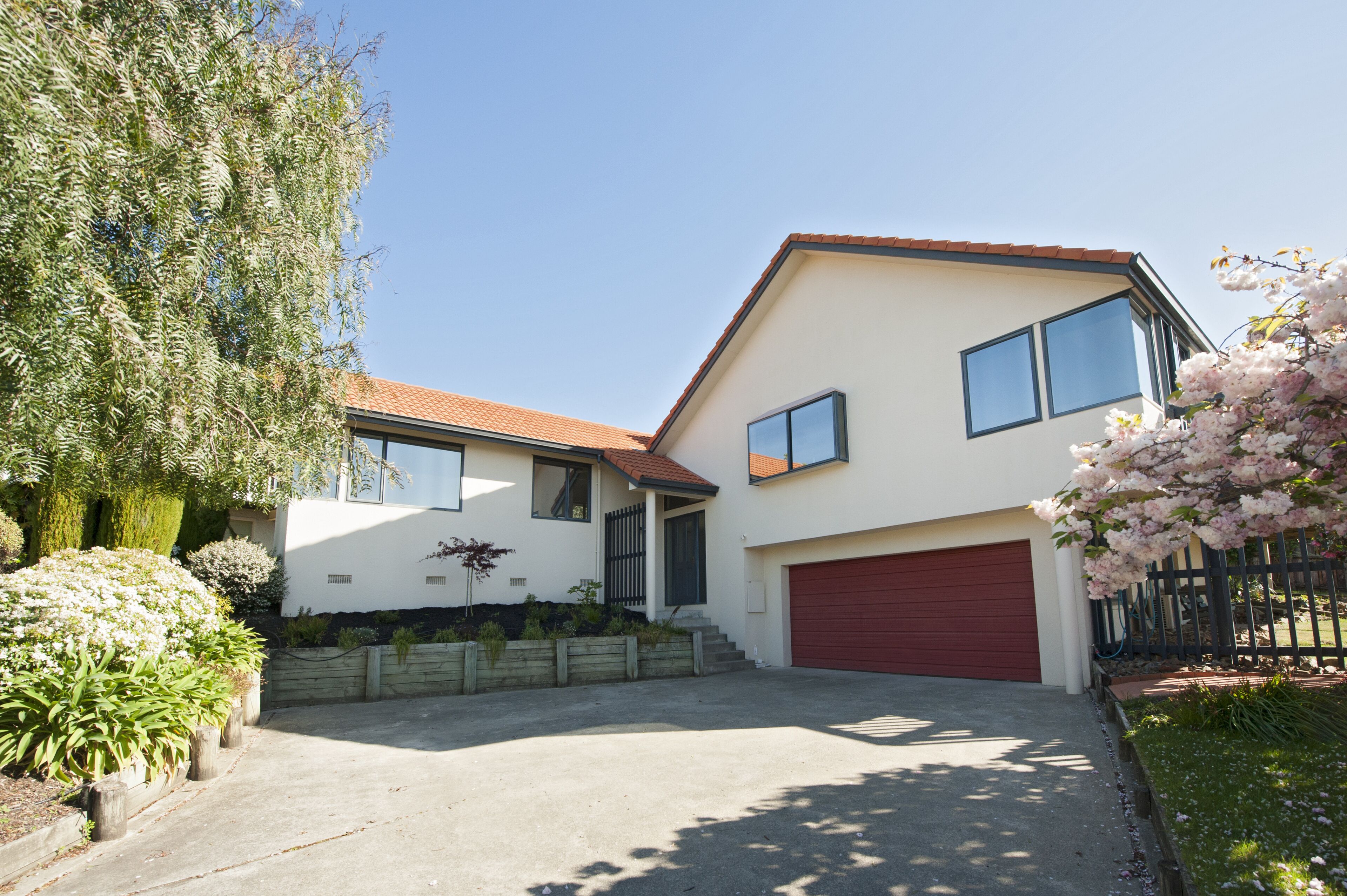 Une maison moderne à deux étages, dotée d’une porte de garage rouge, d’un toit en pente et d’un jardin bien entretenu à l’avant, agrémenté de diverses plantes et d’un arbre.