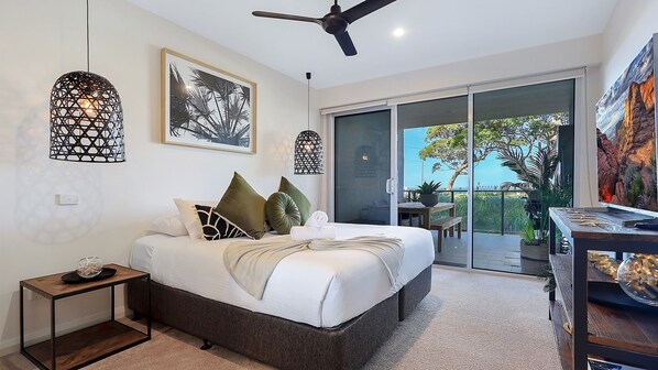 A modern bedroom with a bed, a side table, a ceiling fan, and a large window with a view of greenery.