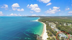 Eine Luftaufnahme einer Küstenlandschaft mit Strand, klarem, blauem Wasser und einer Wohngegend mit Häusern und viel Grün.