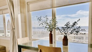 Un comedor con mesa y sillas de vidrio, vista a la playa y una planta en maceta en el alféizar.
