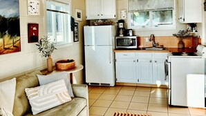 Una cocina compacta con armarios blancos, un refrigerador, un microondas y una estufa. Hay un sofá color beige con cojines y una mesa de centro de madera.