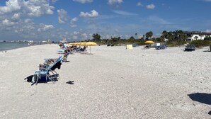 A beach with sun loungers and umbrellas, a clear sky, and a distant view of buildings and palm trees. - Fully Renovated Waterfront Condo on Isla del Sol (St. Petersburg)