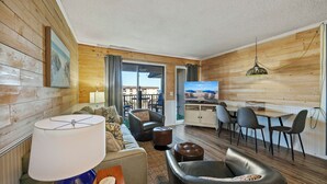 Ein gemütliches Wohnzimmer mit einer Couch, einem Hocker und einem Tisch mit einer Lampe. Es gibt eine Essecke mit Stühlen und einem Fernseher.