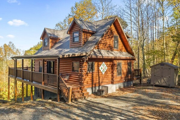 Une cabane en rondins avec un porche, un petit abri de jardin et une forêt en arrière-plan.