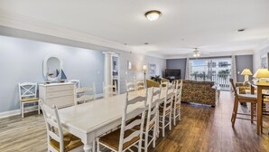 A dining area with a long table and white chairs, a small white dresser, and a television in the background. - Ocean Blvd Luxury Villas 205, Pool & Hot Tub (NORTH MYRTLE BEACH)