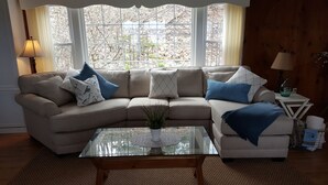 Ein Wohnzimmer mit einem beigefarbenen Ecksofa, einem Glastisch und Blick durch große Fenster.