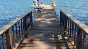 Eine Holzstege, der sich in ruhiges Wasser erstreckt, mit zwei Reihen weißer Liegestühle und Sonnenschirmen am Ende.