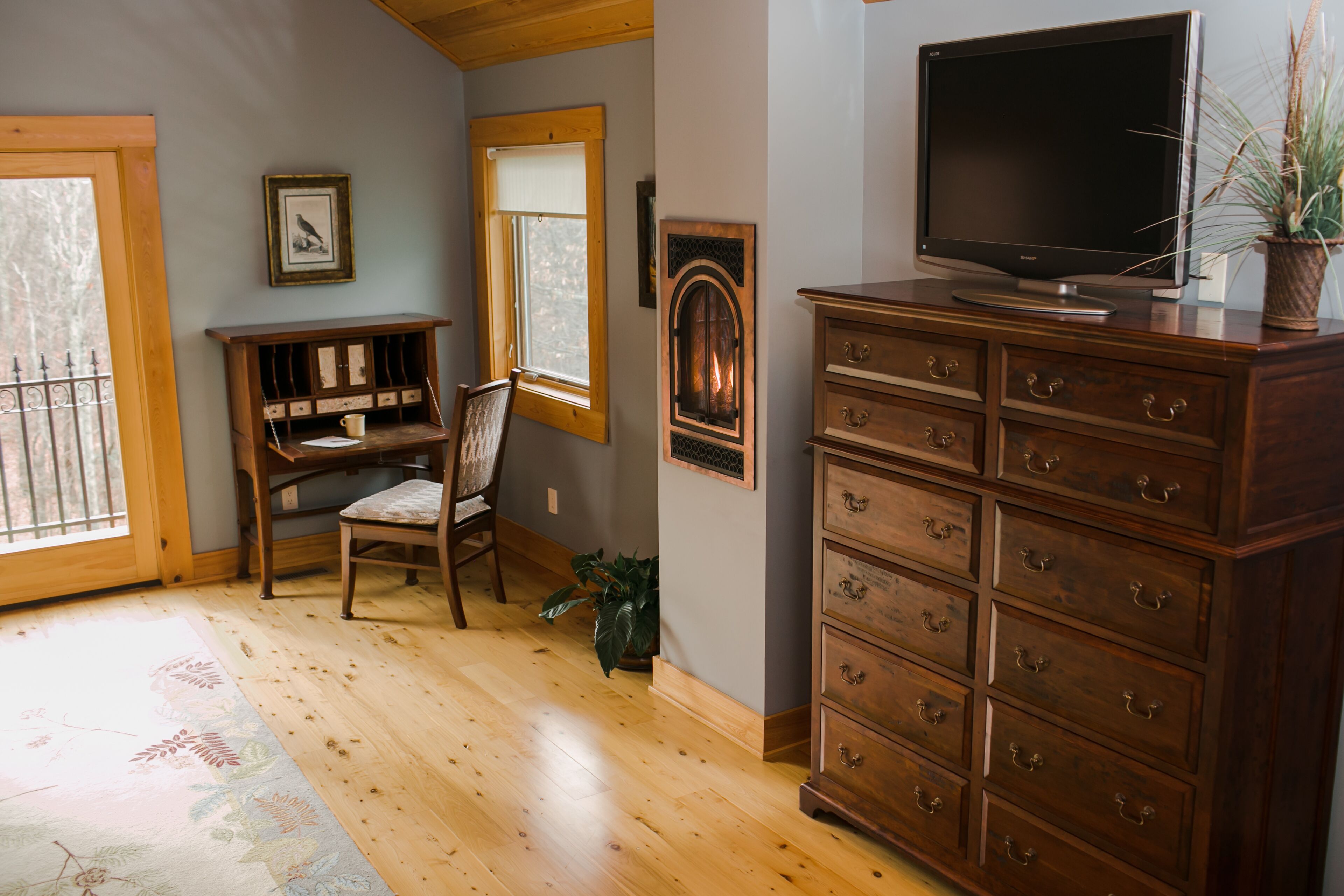 A room with wooden floors, a large wooden dresser with multiple drawers, a television on top, a chair, a desk with a chair, a window, and a framed picture on the wall.