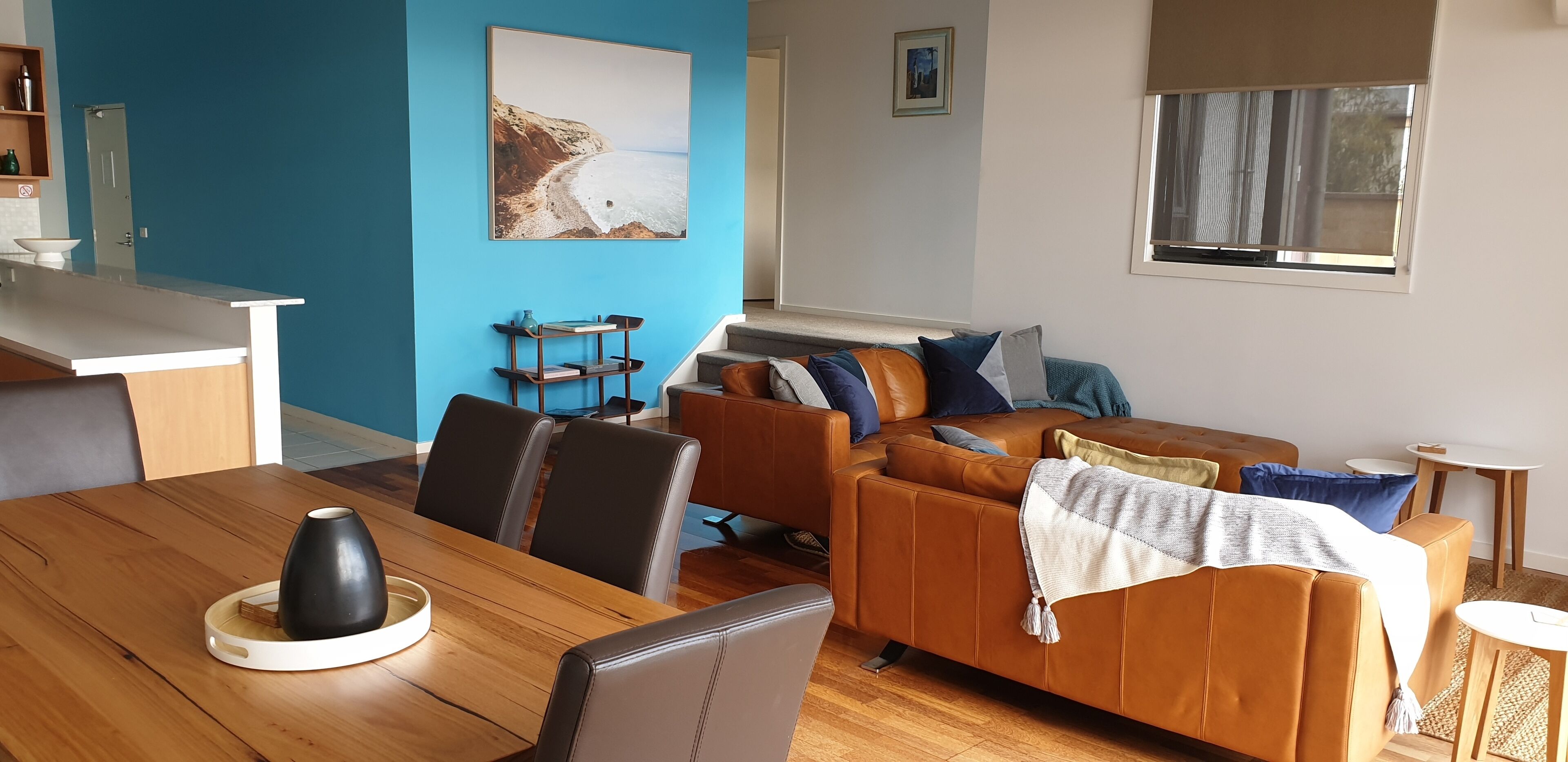 A modern living room with a wooden dining table, leather sofa, and a blue accent wall.