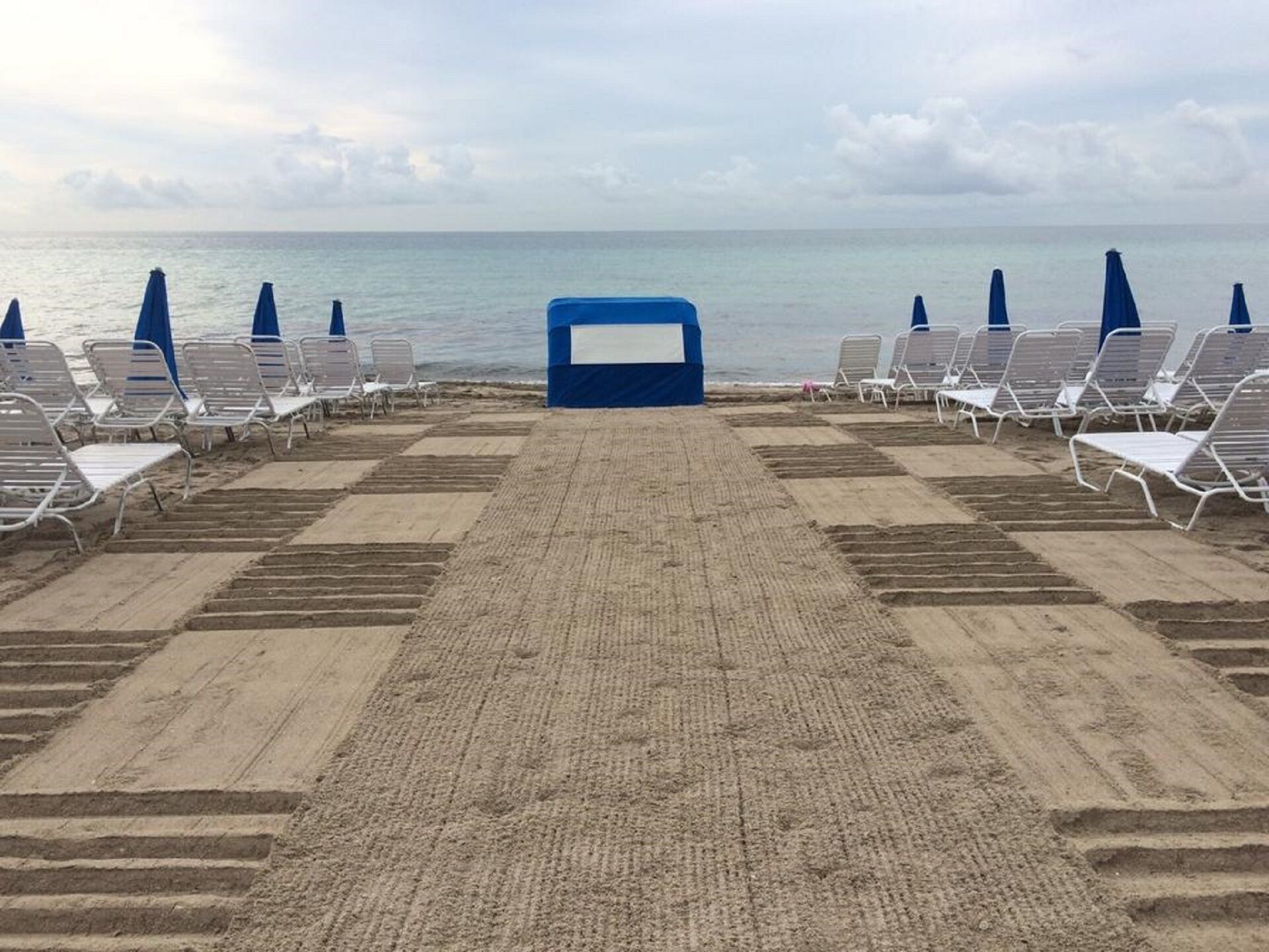 A beach with rows of white lounge chairs and blue umbrellas, leading to a blue building at the water's edge.