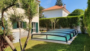 A swimming pool with a glass enclosure, surrounded by a well-maintained lawn and a house with a red-tiled roof. - Lovely House in Lisbon coast For 8 guests (São Domingos de Rana)