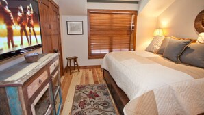 Un dormitorio con una cama, televisión, cómoda de madera, un taburete pequeño, una ventana con persianas y una lámpara.