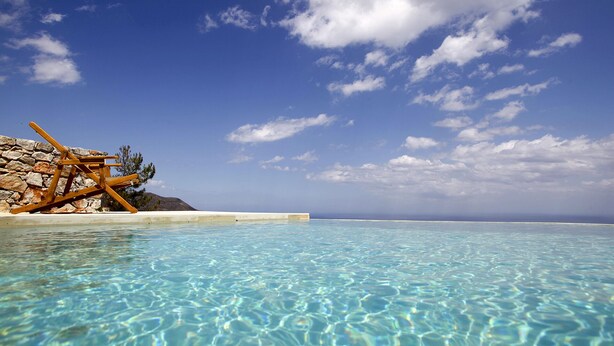 Uma piscina azul cristalina com um muro de pedra e uma estrutura de madeira à esquerda.