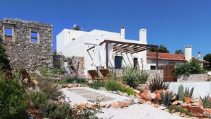 Um edifício de pedra tradicional com uma estrutura moderna em branco, um jardim com diversas plantas e um céu azul límpido.