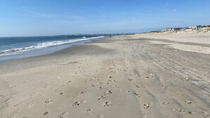 Une vaste plage de sable où l’on peut voir des empreintes de pas menant à l’eau, un ciel dégagé et une côte lointaine parsemée de bâtiments.