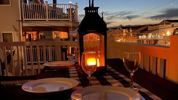 Une table dressée pour deux personnes, avec assiettes, couverts et verres à vin, une lanterne allumée et un balcon donnant sur un quartier résidentiel.