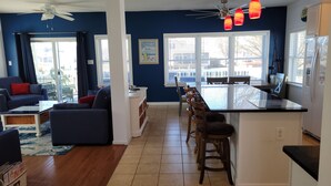 A modern living room with a sofa, a dining area with chairs, a kitchen with a bar, and large windows.