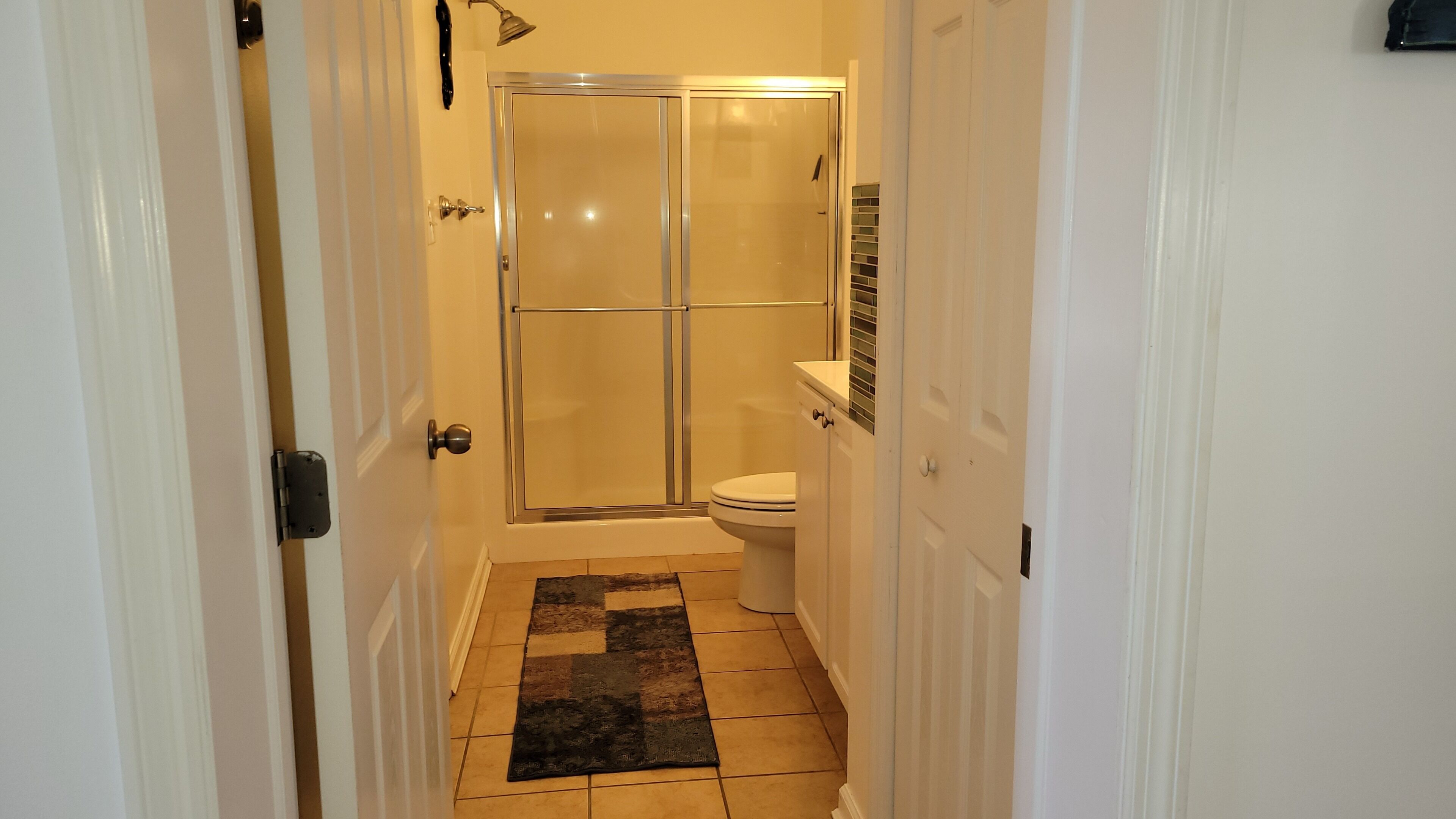 Un pasillo estrecho que conduce a un baño con cabina de ducha de vidrio, un inodoro y suelo de baldosas.