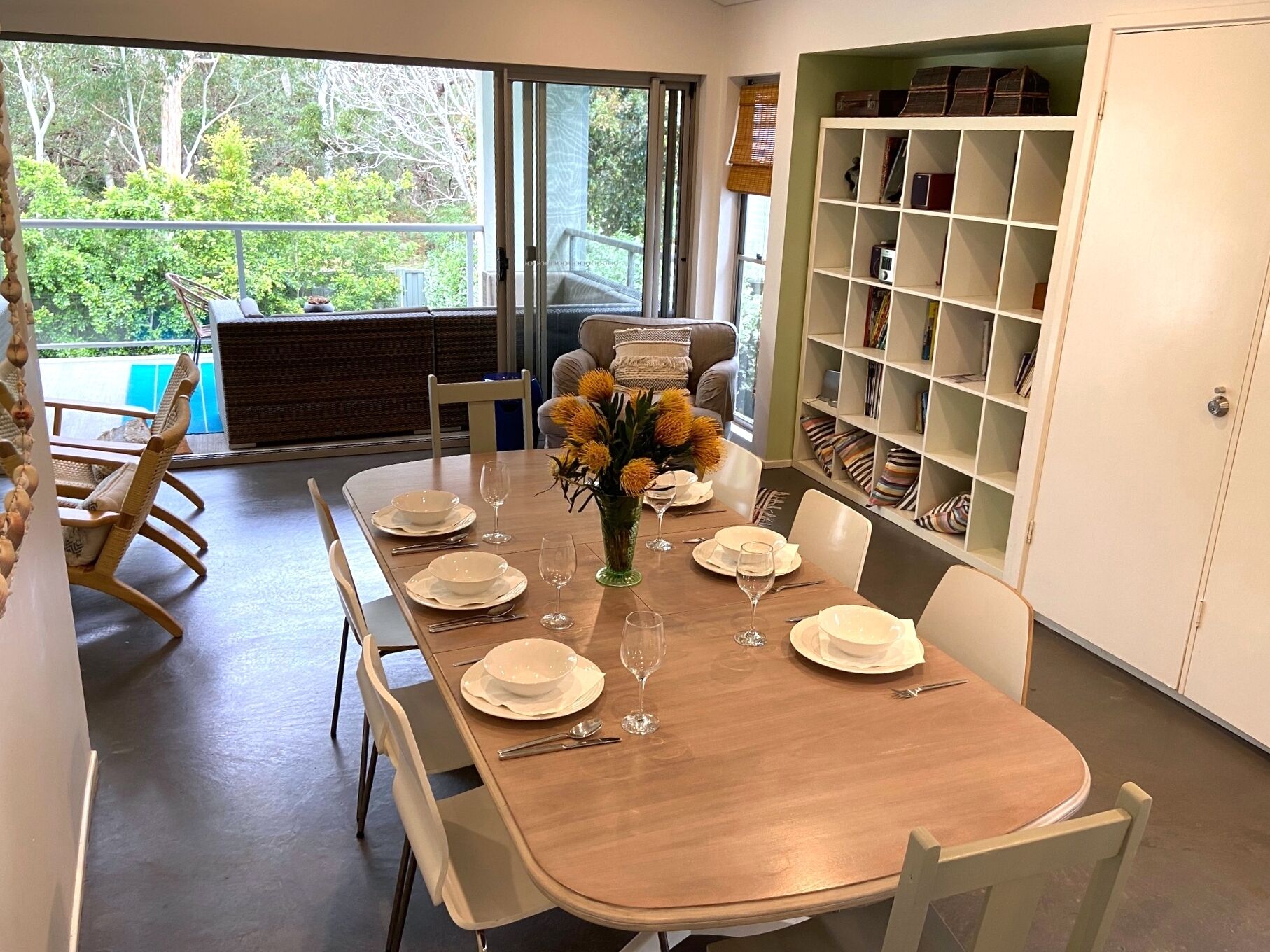 A dining area with a wooden table set for six, surrounded by white chairs. A bookshelf with various items is to the right.