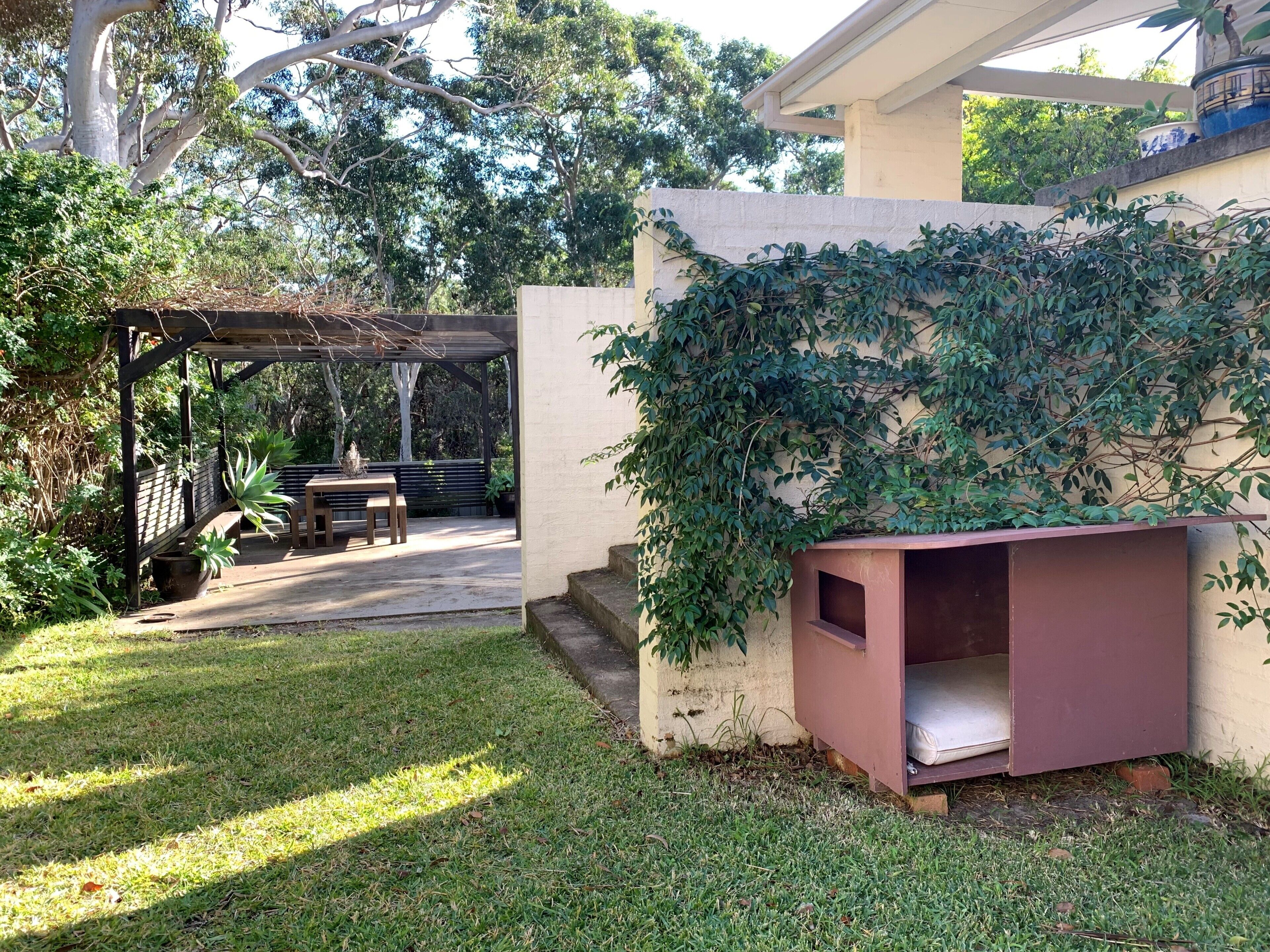A covered outdoor seating area with a table and chairs, surrounded by lush greenery and trees.