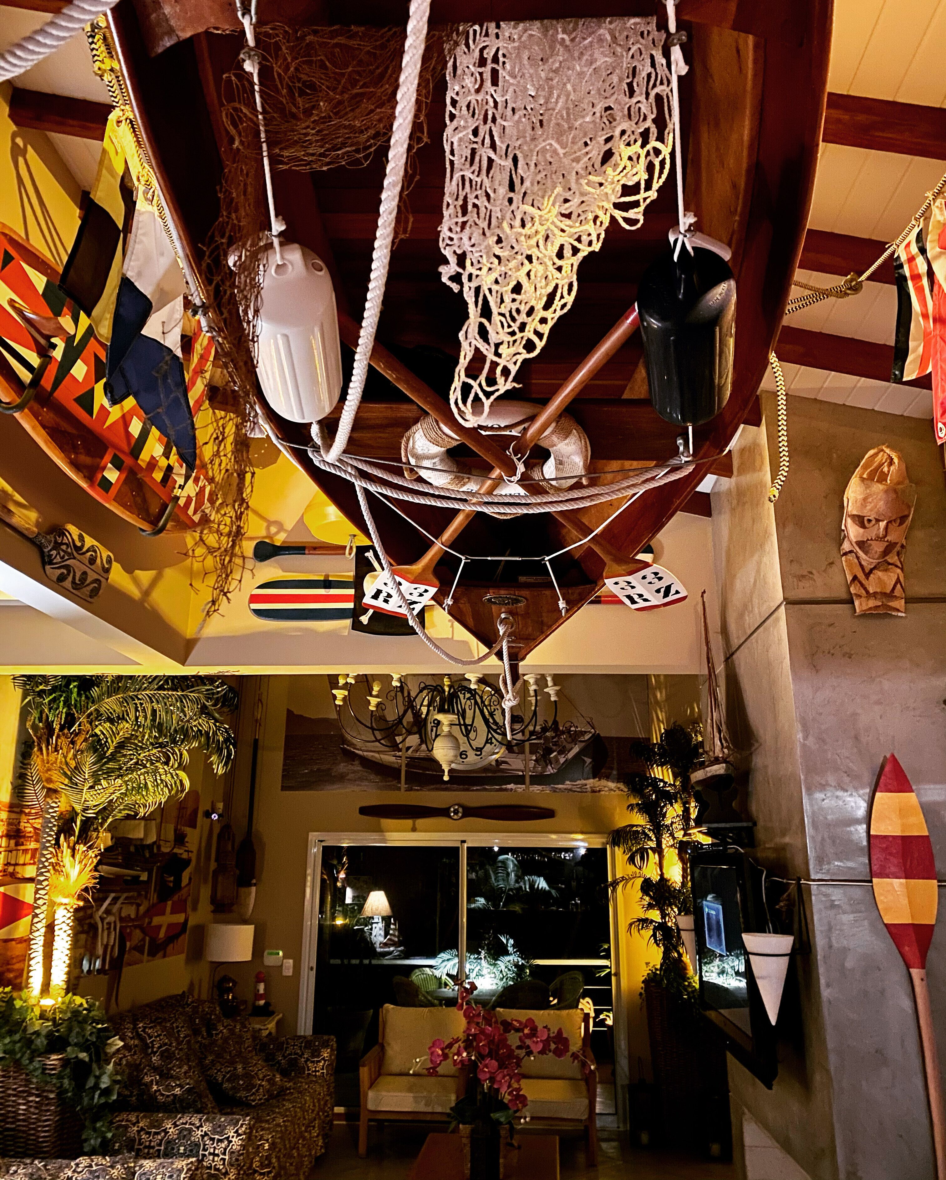 Una stanza con una ventola a soffitto, un lampadario, una tavola da surf e una scultura tiki.