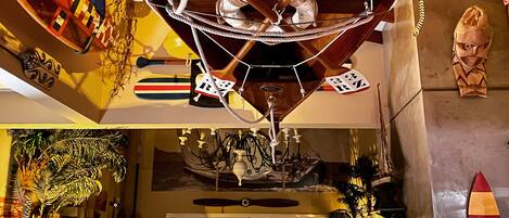 Una stanza con una ventola a soffitto, un lampadario, una tavola da surf e una scultura tiki.