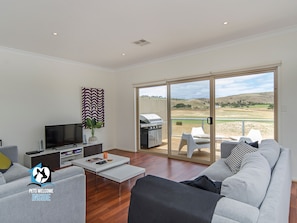 A spacious living room with a flat-screen TV, a coffee table, and a sofa. There is a sliding glass door leading to an outdoor patio with a grill and a view of the countryside.