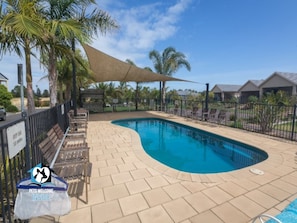 A swimming pool with a curved edge, surrounded by a fence and shaded by a canopy. There are lounge chairs and a sign indicating 'Pets Welcome Inside'.