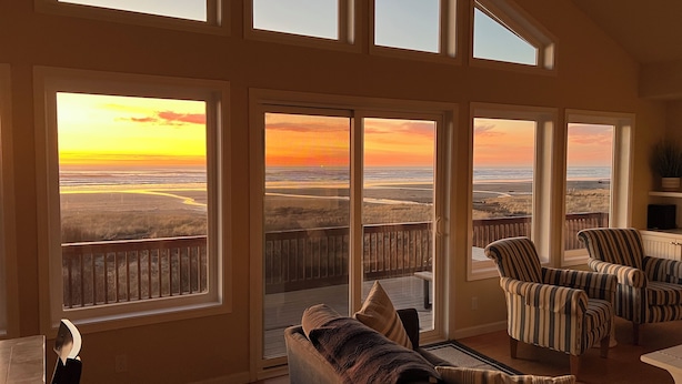 A spacious living room with large windows offering a view of a sunset over a coastal landscape. - Oceanfront Beach House-Nestled on the N. Beach of the Washington Coast (Moclips)