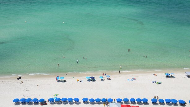 Ein Strand mit klarem türkisblauem Wasser, Menschen beim Schwimmen und Sonnenbaden sowie Reihen blauer Sonnenschirme am Ufer.
