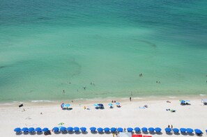 A beach with clear turquoise water, people swimming and sunbathing, and rows of blue umbrellas along the shore.