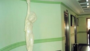 Ein Korridor mit einer weißen Statue auf einer schwarzen Plattform, grünen Wänden und einer Tür mit einem Spiegel.
