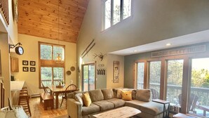 A spacious living area with a large wooden ceiling, a cozy sectional sofa, a dining table with chairs, and a view of the outdoors through large windows. - Luxurious, Secluded, Ski Chalet, w/ Country Decor & Hot Tub Near Okemo (Ludlow)
