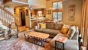 A spacious living area with a large couch, a wooden coffee table, and a staircase leading to an upper level. - Luxurious, Secluded, Ski Chalet, w/ Country Decor & Hot Tub Near Okemo (Ludlow)