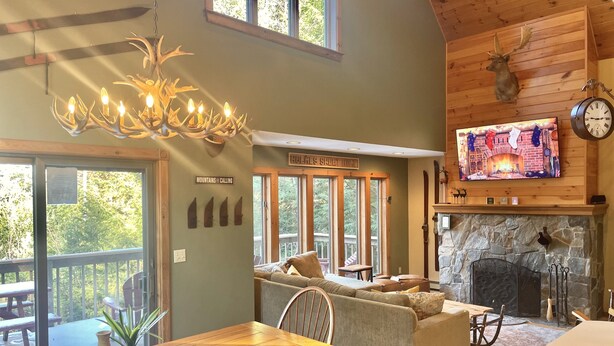 A cozy living room with a fireplace, a wooden ceiling, and a large window overlooking a deck. - Luxurious, Secluded, Ski Chalet, w/ Country Decor & Hot Tub Near Okemo (Ludlow)