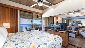 Habitación de hotel con una cama grande, un balcón con vista al océano, una televisión y una zona de estar con sofá y sillones.