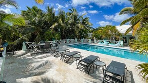 A pool area with lounge chairs and tables, surrounded by palm trees and a clear sky.