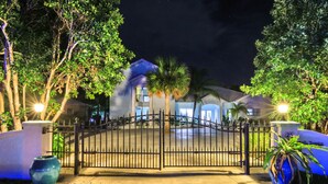 A gated entrance to a property with a well-lit pathway, palm trees, and a large decorative vase.