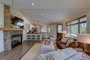 Amplia sala de estar con chimenea, cocina con taburetes de bar, sofá cómodo y ventanas grandes.